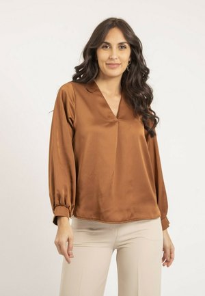 Blouse en satin marron avec un décolleté en V et des manches longues, présentant des poignets froncés et une coupe ample, associée à un pantalon beige clair.