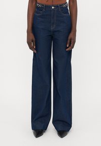 Jean large en denim bleu avec taille haute, présentant des accents en doré sur la ceinture et un design classique à cinq poches.