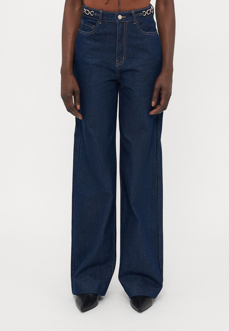 Jean large en denim bleu avec taille haute, présentant des accents en doré sur la ceinture et un design classique à cinq poches.