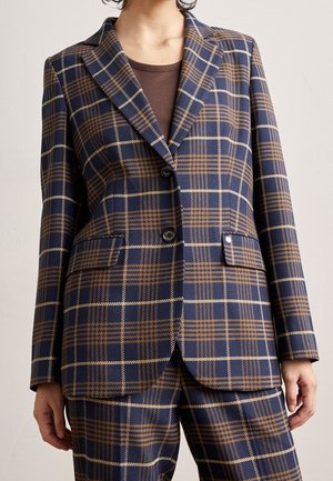 Persoon draagt een marineblauw en bruin geruite blazer met bijpassende broek en een bruin overhemd eronder, staand tegen een effen achtergrond.
