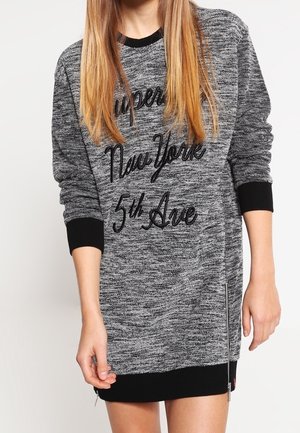 Grijze sweaterjurk met lange mouwen, zwarte manchetten en de tekst "Superdry New York 5th Ave," gedragen door een persoon met steil blond haar.