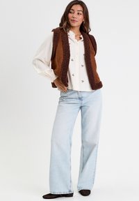 Gilet in suede marrone con foderatura in pelliccia marrone, indossato sopra una blusa chiara. Jeans a gamba larga blu chiaro e scarpe in rete nere completano l'outfit.