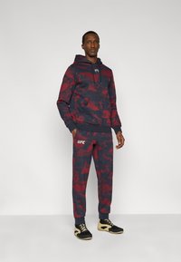 Σετ φούτερ με κουκούλα και παντελόνι σε σχέδιο καμουφλάζ σε ναυτικό μπλε και μπορντό. Διαθέτει ελαστικά μανσέτα, τσέπη καγκουρό και λογότυπο UFC στο παντελόνι.