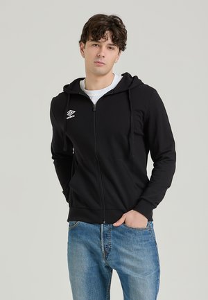 Felpa nera con zip realizzata in tessuto morbido, con un logo bianco sul petto, cappuccio con cordino e tasche a canguro. Indossata con jeans blu.