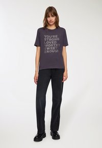 T-shirt grigio scuro con scollo a girocollo che presenta un testo bianco che dice: "SEI FORTE, AMATO, MERITEVOLE E SUFFICIENTEMENTE DOLCE!" Abbinato a jeans neri e stivali neri chunky.