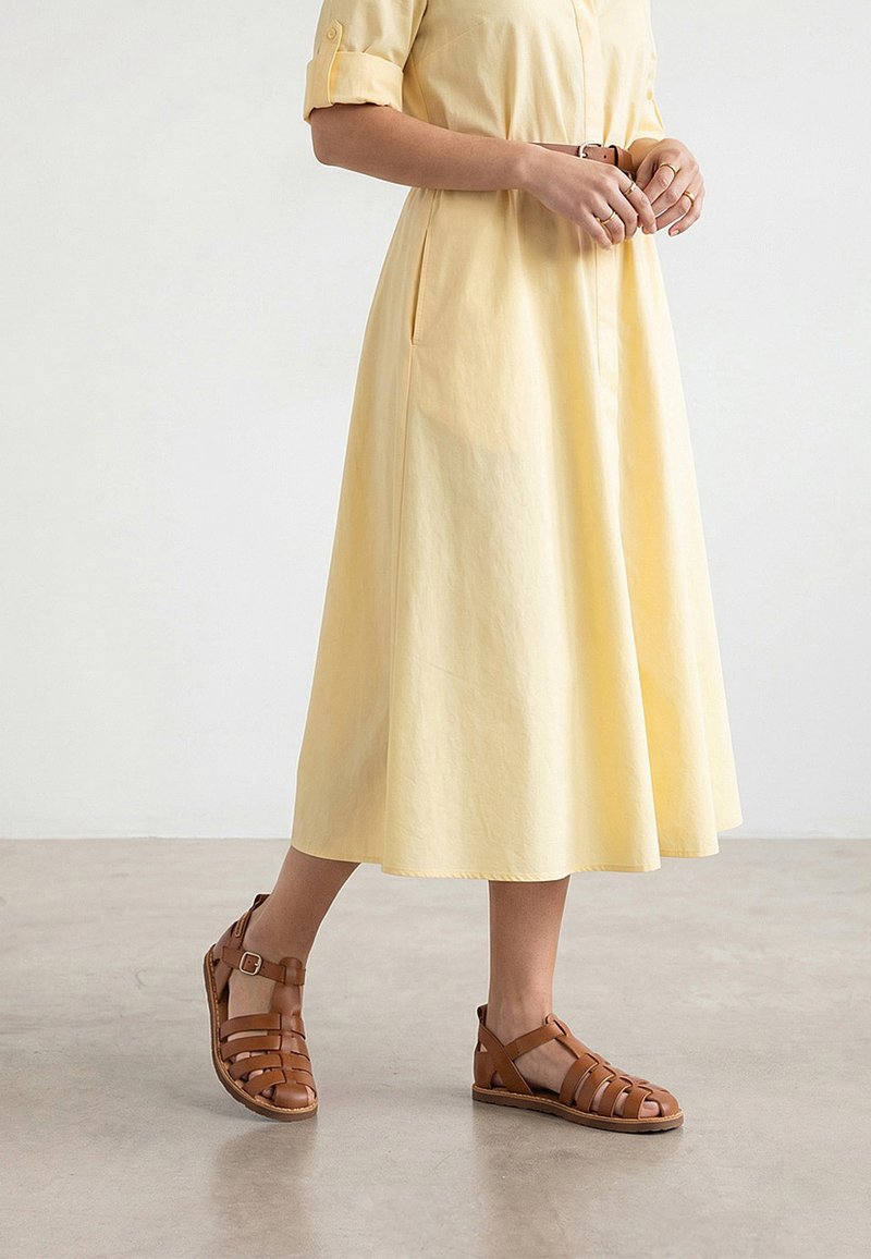 Donna che indossa un vestito midi giallo chiaro con una cintura marrone e sandali piatti marroni con cinturini, in piedi su un pavimento in cemento liscio.