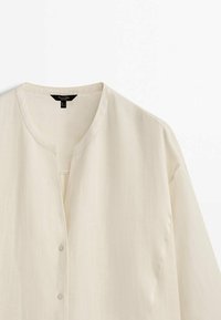Chemise beige clair à boutons avec col rond et manches longues, présentée sur fond blanc.