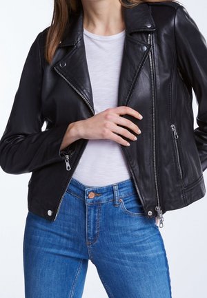 Femme portant une veste en cuir noire par-dessus une chemise blanche et un jean bleu, ajustant la fermeture éclair de la veste avec sa main droite.