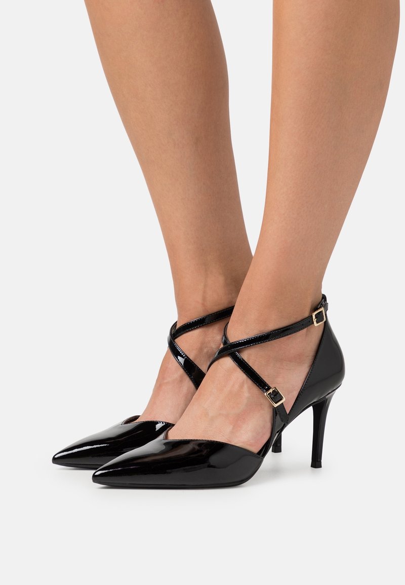 Escarpins à talons aiguilles en cuir verni noir, à bout pointu, avec des brides croisées à la cheville et de petites boucles dorées, portés pieds nus sur un fond blanc.