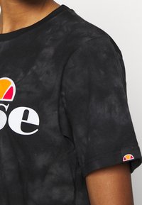 Torso superior vestido con una camiseta negra de tie-dye con un logo impreso colorido y una pequeña etiqueta de logo a juego en la manga.