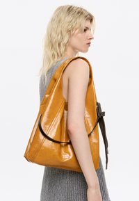 Mujer rubia con pelo ondulado lleva un vestido de punto gris sin mangas y sostiene un grande bolso amarillo mostaza brillante en su hombro.