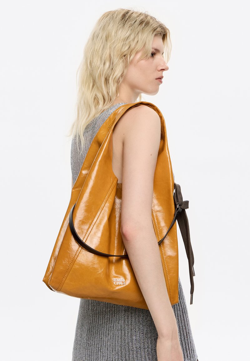 Mujer rubia con pelo ondulado lleva un vestido de punto gris sin mangas y sostiene un grande bolso amarillo mostaza brillante en su hombro.