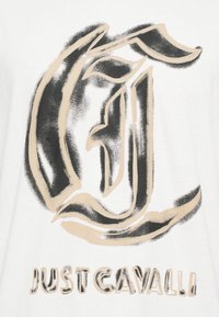 Balta marškinėliai su dideliu juodu ir smėlio spalvos grafiniu logotipu, kurio pavidalu rašoma „C“ kursyvu, o po juo yra užrašas „JUST CAVALLI“ ryškiu šriftu.