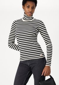Maglione a dolcevita a righe bianche e nere con texture a coste, silhouette aderente e logo CK sull'orlo, abbinato a jeans scuri e una borsa nera.