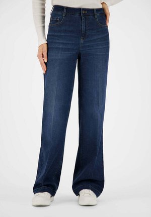 Frau trägt dunkelblaue Jeans mit weitem Bein und hoher Taille, kombiniert mit weißen Sneakers und einem weißen, gerippten Langarmshirt.