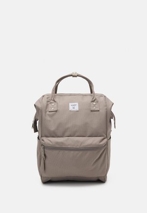 Sac à dos beige de forme carrée avec doubles poignées, poche zippée à l'avant, poches latérales et une petite étiquette de marque blanche centrée près du haut.