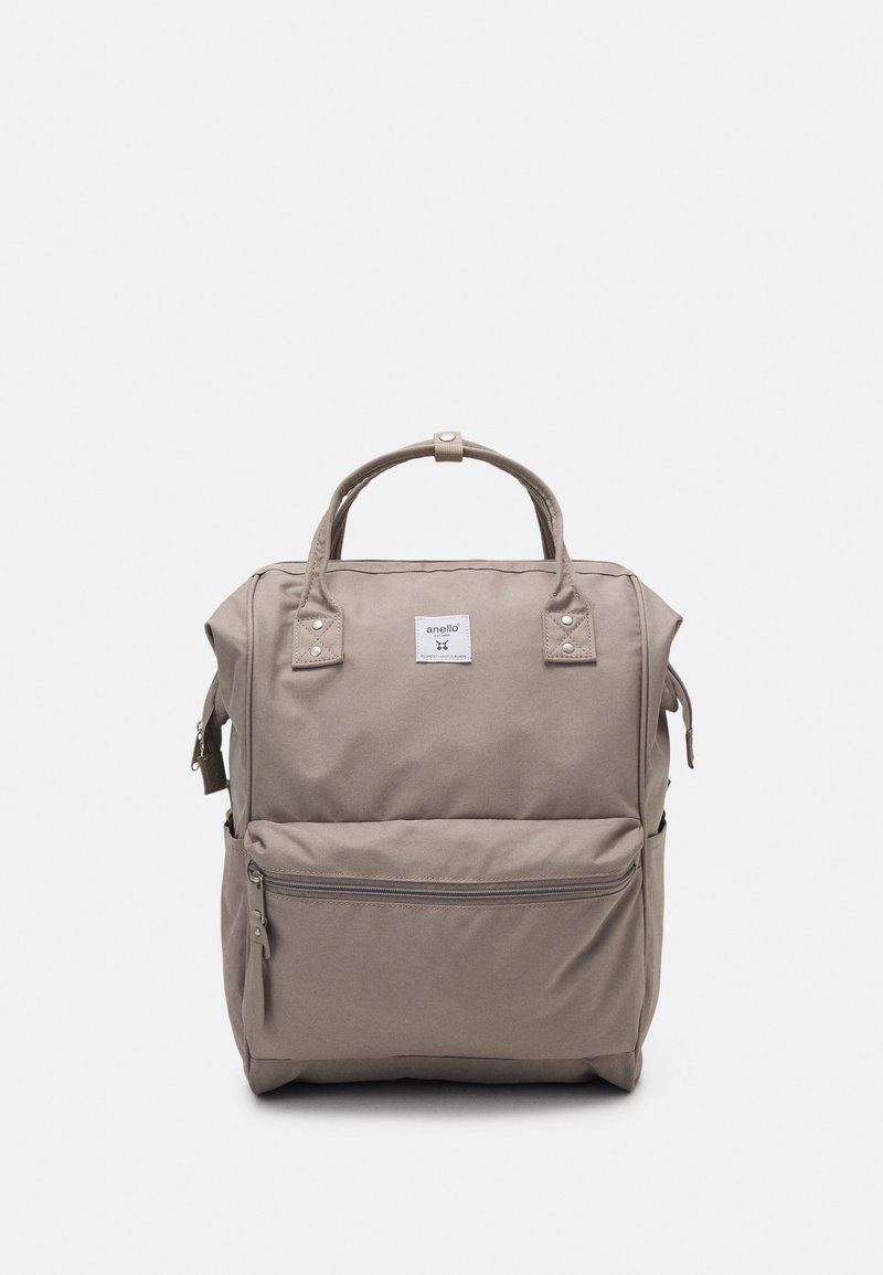 Sac à dos beige de forme carrée avec doubles poignées, poche zippée à l'avant, poches latérales et une petite étiquette de marque blanche centrée près du haut.