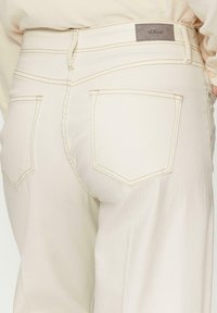 Pantalons taille haute beiges en tissu doux, avec des coutures contrastantes, deux poches arrière et une étiquette rectangulaire à la taille.