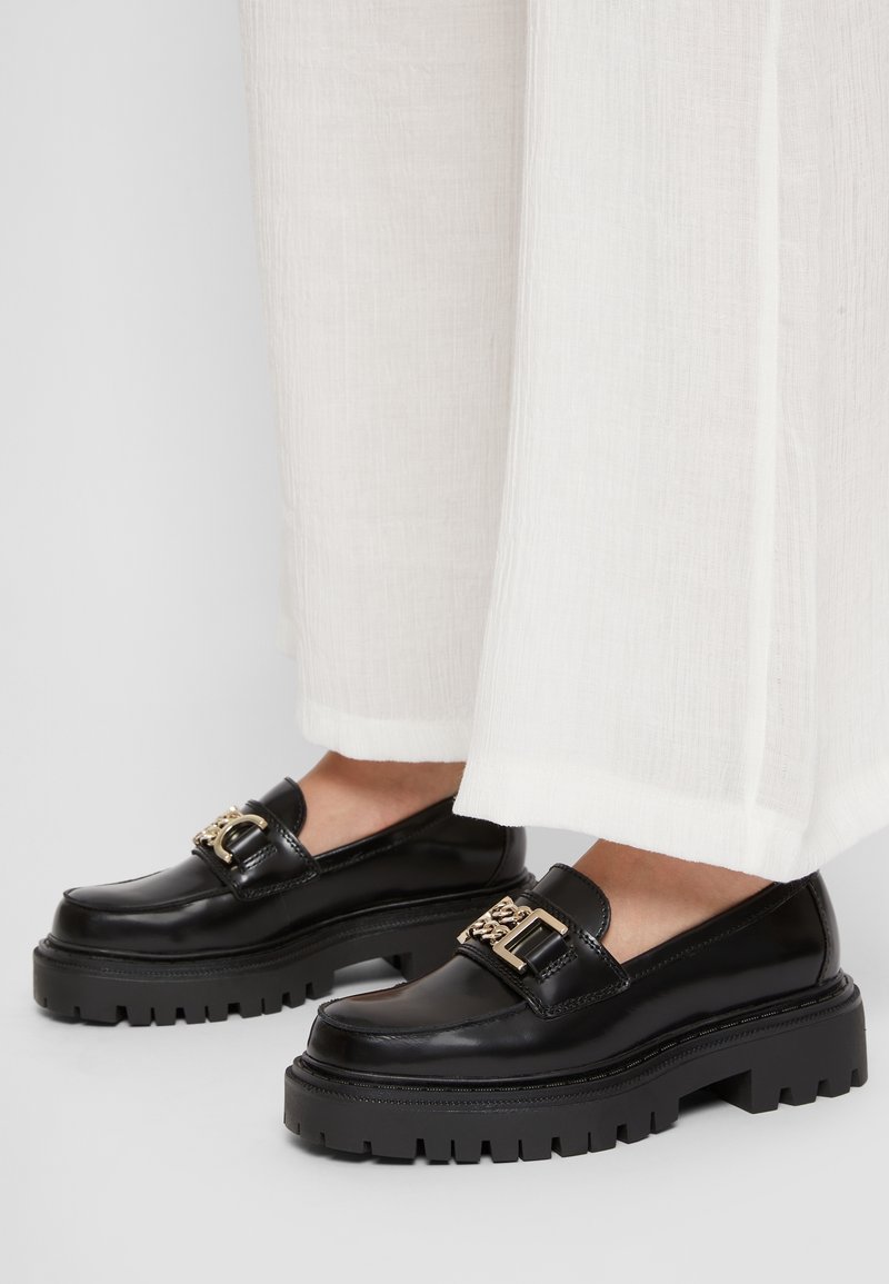 Mocassins à plateforme noire épaisses avec détail chaîne dorée, portés avec un pantalon blanc à jambes larges.