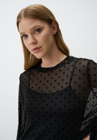 Schwarze durchsichtige Bluse mit Punkten, die lange Ärmel und einen runden Halsausschnitt hat. Der Stoff wirkt leicht und strukturiert.