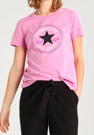 Persoon gekleed in een roze Converse All Star t-shirt en zwarte broek met een strik in de taille, staand met één hand in de zak.