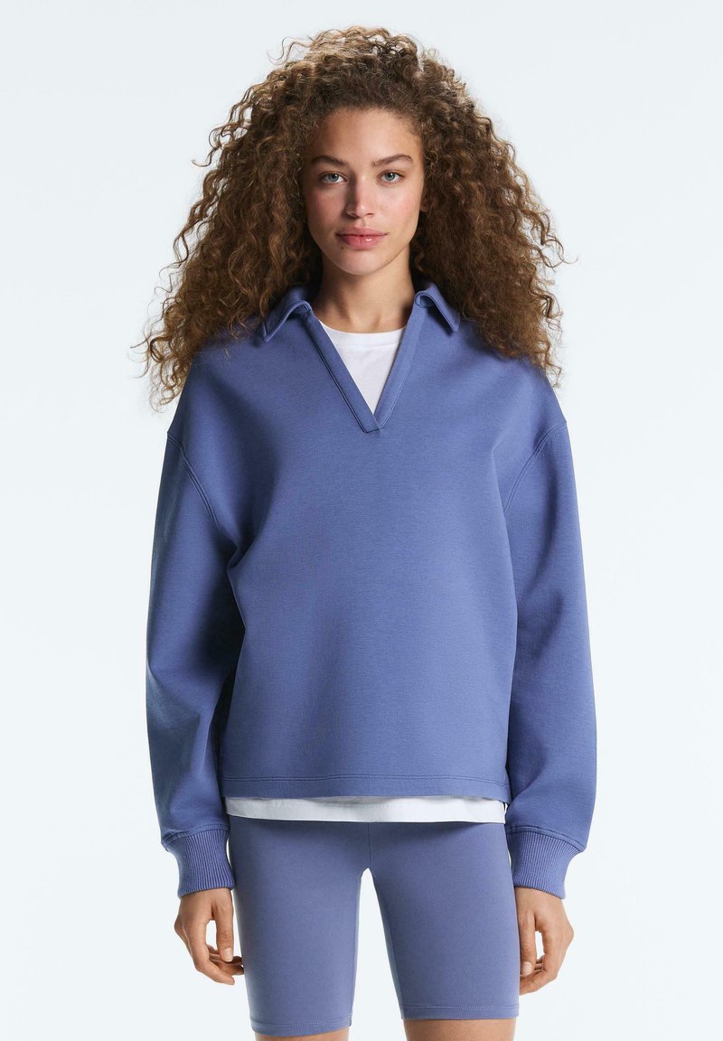 Femme aux cheveux bouclés portant un sweat-shirt bleu oversize avec col et un short de vélo bleu assorti, debout devant un fond uni.