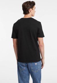 T-shirt noir en coton à manches courtes et col rond, vu de dos. Associé à un jean en denim bleu avec un patch logo triangulaire.