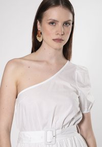 Vestido blanco de un solo hombro con manga abullonada, cintura ceñida con un cinturón blanco grueso, delicados detalles de ojales en el dobladillo, textura suave.