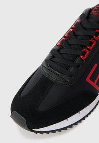 Schwarze Sneaker aus einer Kombination aus glattem Synthetikmaterial und strukturiertem Wildleder. Rote Akzente an den Schnürsenkeln und im Branding; weiße Sohle mit schwarzem Profil.
