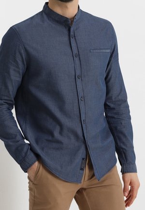 Muž v modré košili na knoflíky s mandarínským límečkem a béžovými kalhotami stojí s jednou rukou v kapse před jednoduchým pozadím.