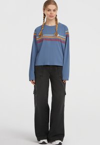 Blue long-sleeve shirt with horizontal stripes in red, yellow, and orange. Paired with loose black cargo pants. White sneakers visible.