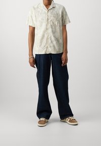 Korte mouwen, lichtgekleurde shirt met een bladerenpatroon, gecombineerd met losse donkerdenim broek en bruine sneakers met witte accenten.