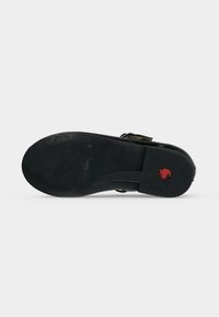 Semelle de chaussure noire avec un petit logo circulaire rouge près du talon, nom de marque embossé visible et une boucle de sangle sur le côté.