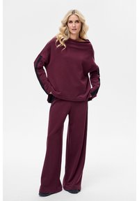 Bordowa, oversize bluza z dekoltem typu łódka i teksturowanymi czarnymi panelami po bokach, zestawiona z pasującymi szerokimi spodniami z miękkiego materiału.