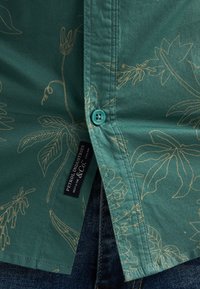 Chemise verte avec motifs floraux beiges, bouton turquoise, fente latérale et étiquette noire sur un jean en denim bleu.