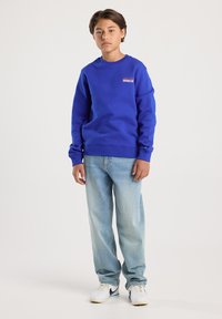 Adolescente che indossa un felpa blu con la scritta "Brooklyn", jeans azzurri e sneakers bianche, in piedi contro uno sfondo chiaro e semplice.