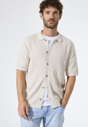 Chemise beige à manches courtes avec boutons, en tissu texturé, présentant un col classique et une coupe décontractée, portée par-dessus un t-shirt blanc.