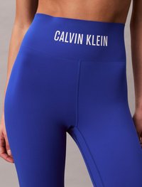 Modre legice visoke v pasu, izdelane iz raztegljivega materiala, z belim logotipom "CALVIN KLEIN" na pasu in gladko, brezšivno teksturo.