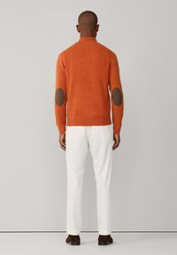 Maglione arancione con toppe marroni sui gomiti, design aderente, polsini a costine e collo alto, abbinato a pantaloni crema e scarpe scure.