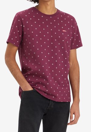 Homme portant un T-shirt bordeaux à manches courtes avec de petits motifs losanges blancs et un pantalon noir, main dans la poche.
