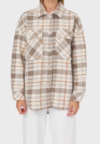 Chemise à carreaux à boutonner en beige, marron et blanc. Fabriquée en tissu doux, avec deux poches poitrine, un col et de longues manches.
