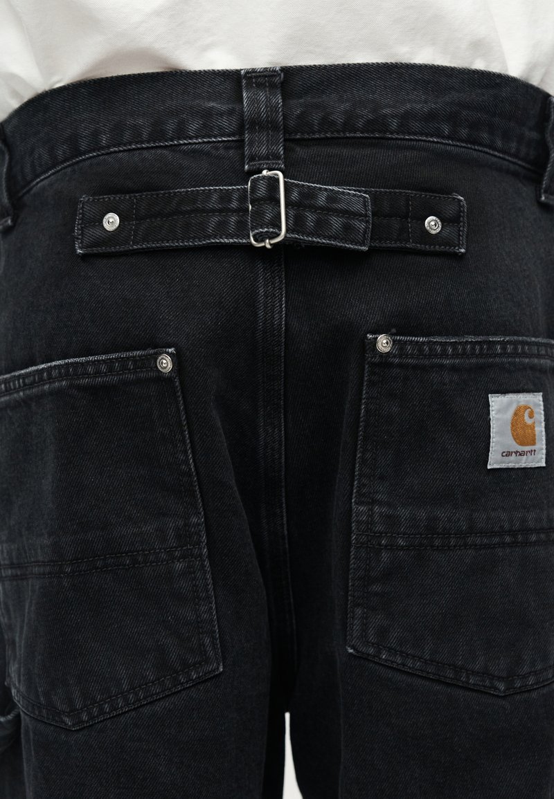 Vista posteriore di jeans neri con cinturino regolabile con fibbia e grandi tasche quadrate, una delle quali presenta una toppa con il logo Carhartt arancione.