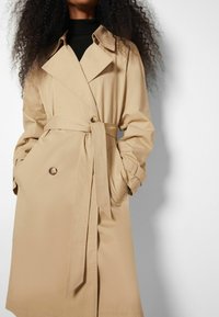 Manteau beige à double boutonnage, ceinture à nouer, accents de boutons et texture de tissu lisse et soyeuse.
