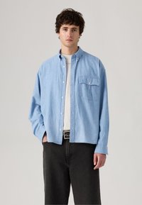 Lysblå denimskjorte med knap-front, enkelt brystlomme og langeærmer. Matchet med sorte bukser og et sort bælte.