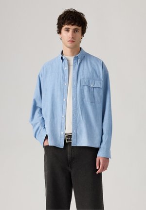 Light blue denim shirt with button front, single chest pocket, and long sleeves. Paired with black pants and a black belt.