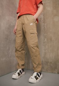 Beige cargobyxor med elastisk midja och sidofickor, med en vit Nike-logotyp; bärs med vita sneakers och en orange t-shirt.