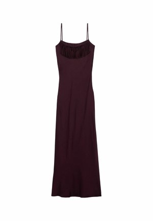 Donna che indossa un vestito lungo viola scuro senza maniche con dettagli di frange, cintura nera e tacchi dorati con cinturini, in piedi su uno sfondo chiaro.