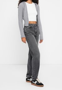 Femme portant un crop top blanc, un sweat à capuche gris en maille torsadée, un jean noir délavé taille haute et des baskets noires avec des touches blanches.