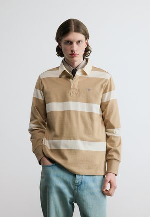 Junger Mann mit schulterlangem Haar trägt ein beige-weiß gestreiftes Langarm-Poloshirt und hellblaue Jeans, posiert vor einem einfarbigen Hintergrund.