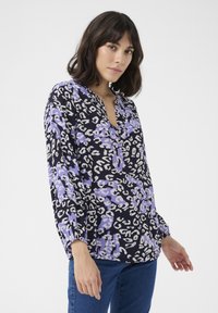 Blouse à manches longues en bleu marine foncé avec un motif coloré de léopard dans des teintes de violet, blanc et gris. Dispose d'un col en V et d'une coupe ample.